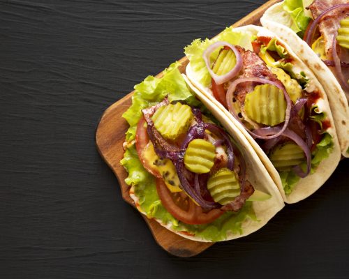 Homemade Bacon Cheeseburger Tacos on a rustic wooden board on a black surface, top view. Flat lay, overhead, from above. Copy space.